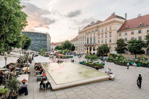 The bustling main courtyard of the Museum Quarter at sunset, with a reflecting pool and visitors sitting nearby. Historic buildings and trees contribute to the peaceful atmosphere.