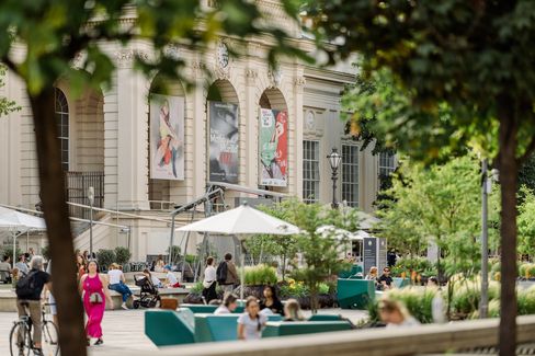 People sit and walk across the green courtyard of the MQ in front of the Tanzquartier with large windows and posters on the facade.