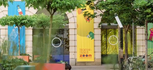 Courtyard with trees, colorful banners on a building, and bicycles next to an entrance with round windows