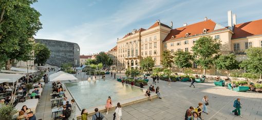 Großer Platz mit langem Wasserbecken, umgeben von historischen Gebäuden und Bäumen, mit vielen Menschen, die sitzen, gehen und sich unterhalten