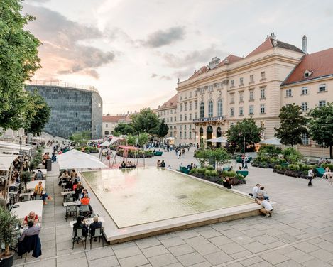 The bustling main courtyard of the Museum Quarter at sunset, with a reflecting pool and visitors sitting nearby. Historic buildings and trees contribute to the peaceful atmosphere.