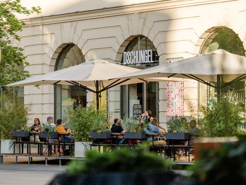 Gäste sitzen im Außenbereich des Restaurants Dschungel Wien im MuseumsQuartier unter großen Sonnenschirmen vor historischer Architektur.