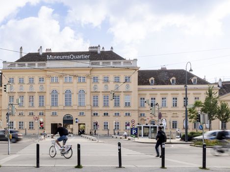 Blick auf das MuseumsQuartier mit einem großen historischen Gebäude, mehreren Personen, einem Radfahrer und Autos auf der Straße davor