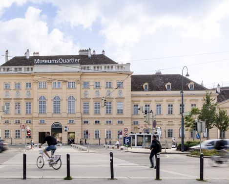 Blick auf das MuseumsQuartier mit einem großen historischen Gebäude, mehreren Personen, einem Radfahrer und Autos auf der Straße davor