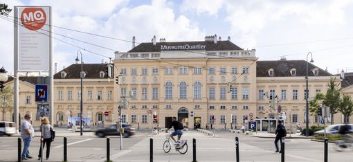Blick auf das MuseumsQuartier mit einem großen historischen Gebäude, mehreren Personen, einem Radfahrer und Autos auf der Straße davor