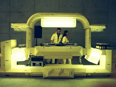 Beleuchtetes DJ-Pult in futuristischem Design mit zwei DJs hinter den Turntables. Die Installation ist aus hellen, geometrischen Elementen aufgebaut und steht in einem großen Betonraum, seitlich sind Getränke und Gläser bereitgestellt.