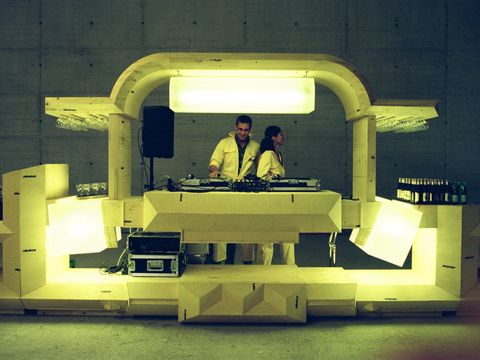 Illuminated futuristic DJ booth with two DJs behind the turntables. The installation is made of light-coloured geometric elements and is set within a large concrete space, with drinks and glasses arranged on either side.