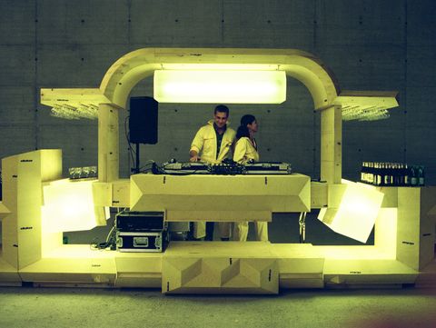 Illuminated futuristic DJ booth with two DJs behind the turntables. The installation is made of light-coloured geometric elements and is set within a large concrete space, with drinks and glasses arranged on either side.