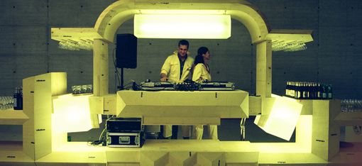 Illuminated futuristic DJ booth with two DJs behind the turntables. The installation is made of light-coloured geometric elements and is set within a large concrete space, with drinks and glasses arranged on either side.