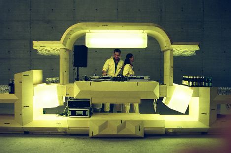 Illuminated futuristic DJ booth with two DJs behind the turntables. The installation is made of light-coloured geometric elements and is set within a large concrete space, with drinks and glasses arranged on either side.
