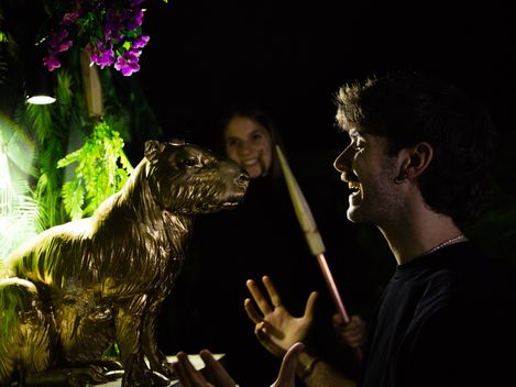 Two people stand in front of a golden animal figure in a lit jungle setting and interact with it.