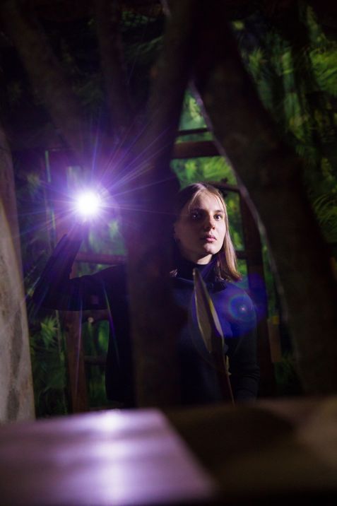 A person stands among branches in a dark room, holding a flashlight that illuminates their face.