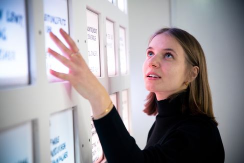 A person gestures in front of a wall of red-lit text panels in an interior space.