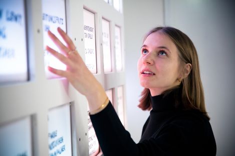 A person gestures in front of a wall of red-lit text panels in an interior space.