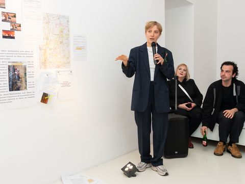 Person mit Mikrofon steht vor einer weißen Wand mit angehefteten Texten und Bildern, zwei weitere Personen sitzen daneben auf dem Boden