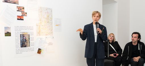 Person holding a microphone stands in front of a white wall with pinned texts and images, two other people sit on the floor nearby