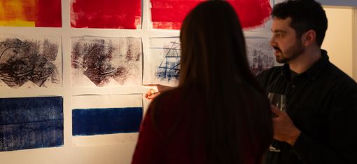 Zwei Personen betrachten eine Wand mit mehreren abstrakten Farb- und Linienzeichnungen in Rot, Blau und Schwarz.