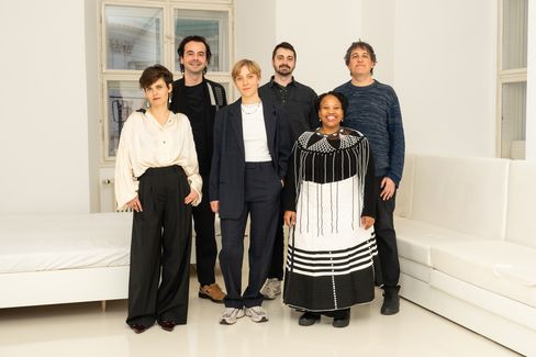 Six people stand in a bright room with white walls and white seating, wearing various casual and formal clothing.