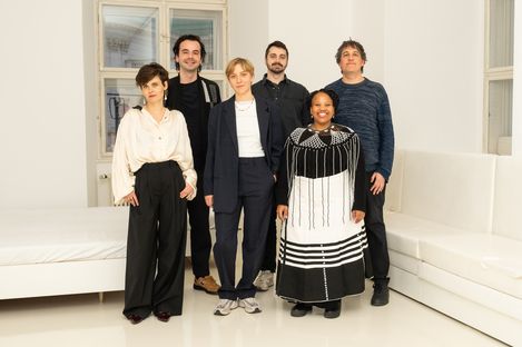 Six people stand in a bright room with white walls and white seating, wearing various casual and formal clothing.