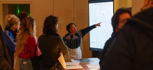 A woman points to a screen, surrounded by people watching her.