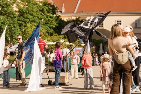 Kinder und Erwachsene stehen auf einem Platz, einige Kinder halten große Fahnen, im Hintergrund Gebäude und Bäume