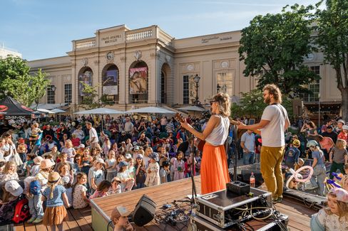 Two musicians on stage in front of a large audience in front of a historic building with trees and parasols