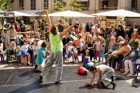 Two children and an adult perform movements on a small stage in front of a seated audience, while market stalls and trees can be seen in the background.
