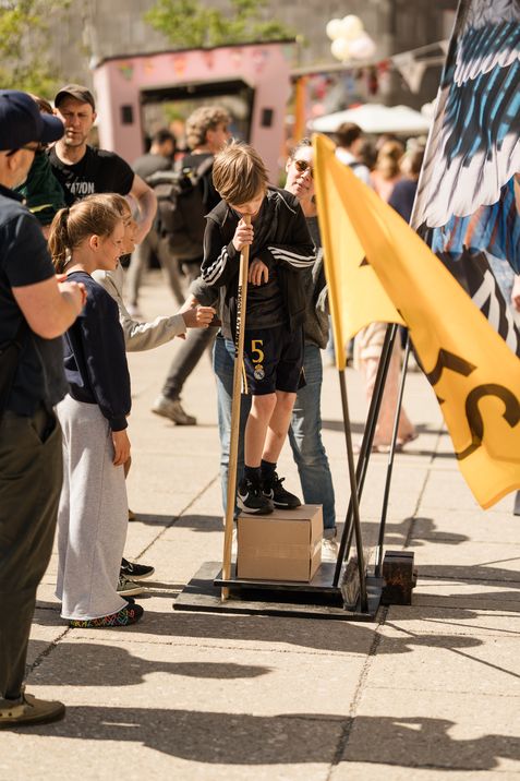Kind steht auf einem Holzbrett mit zwei Stöcken und wird von mehreren Personen umgeben, im Hintergrund ein gelbes Banner und weitere Menschen auf einem Platz