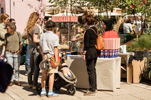 Several people are standing at an outdoor popcorn stand, one person is holding a skateboard, another is pushing a stroller.