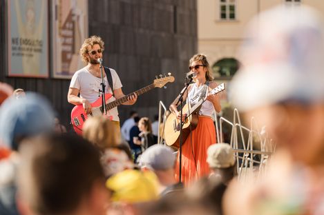 Two people play guitar and bass on an outdoor stage in front of an audience.