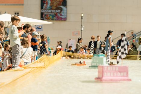Children stand at a course marked with barrier tape and watch small vehicles driving on a track between checkered finish flags.