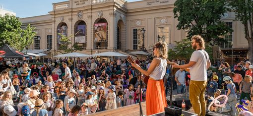 Zwei Musiker auf einer Bühne vor einem großen Publikum vor einem historischen Gebäude mit Bäumen und Sonnenschirmen