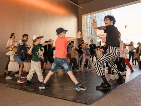 Kinder und ein Erwachsener stehen auf einer schwarzen Matte und machen synchron eine Bewegung mit ausgestrecktem Arm und gebeugtem Knie.