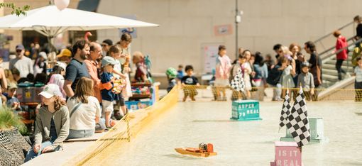 Children and adults watch small boats in a shallow water basin enclosed by a net.