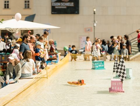 Children and adults watch small boats in a shallow water basin enclosed by a net.