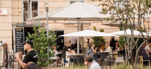 Large outdoor seating area at MuseumsQuartier Vienna with many visitors at tables and standing areas under sun umbrellas.
