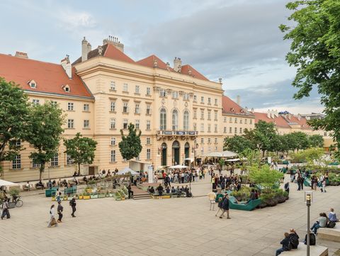Großer Platz mit Bäumen, Sitzgelegenheiten und Menschen vor einem historischen Gebäude mit roten Dächern unter bewölktem Himmel