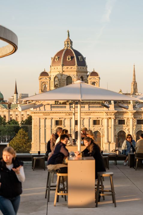 Guests sit at high tables on the MQ Libelle rooftop terrace at MuseumsQuartier Vienna, with the Kunsthistorisches Museum in the evening light in the background.