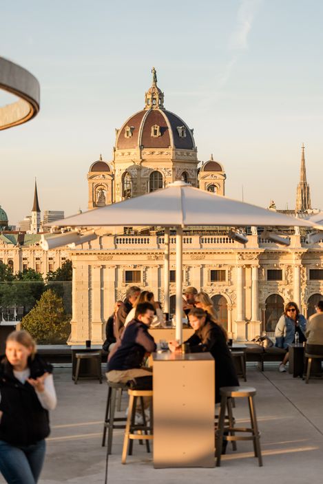Guests sit at high tables on the MQ Libelle rooftop terrace at MuseumsQuartier Vienna, with the Kunsthistorisches Museum in the evening light in the background.