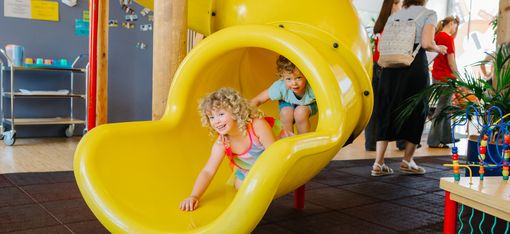 Zwei kleine Kinder rutschen fröhlich aus einer leuchtend gelben Indoor-Rutsche.
