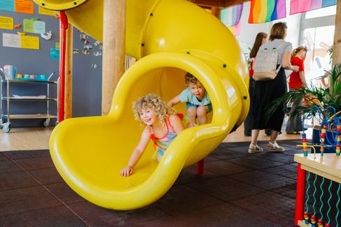 Two young children are happily sliding out of a bright yellow indoor slide.