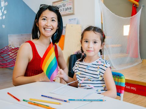 Frau und Mädchen sitzen an einem Tisch in der WIENXTRA-Kinderinfo, lächeln in die Kamera und halten gemeinsam eine Regenbogenflagge; vor ihnen liegen Papier und bunte Stifte.