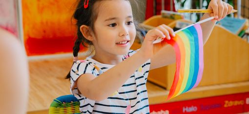 Ein Mädchen sitzt an einem Tisch in der WIENXTRA-Kinderinfo, lächelt und hält eine Regenbogenflagge; vor ihm liegen Papier und bunte Stifte.