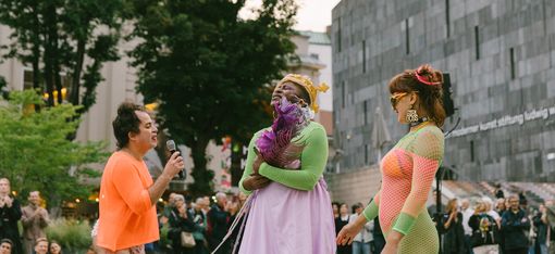 Drei Personen in auffälliger Kleidung stehen vor einer Menschenmenge im Freien, eine Person hält ein Mikrofon.