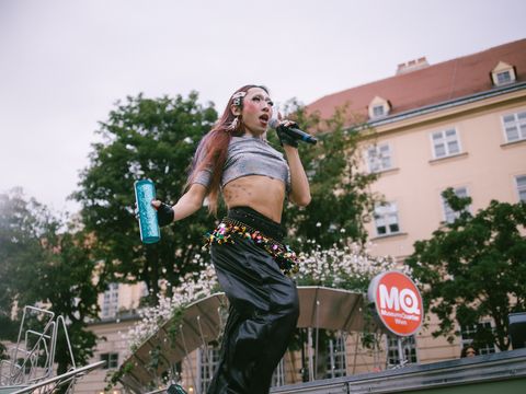 Person with long hair wearing a crop top holding a microphone and a water bottle on a stage in front of a building with an MQ logo
