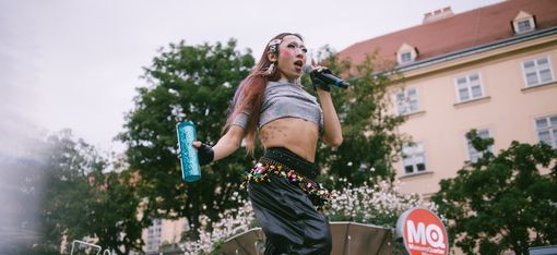 Person with long hair wearing a crop top holding a microphone and a water bottle on a stage in front of a building with an MQ logo