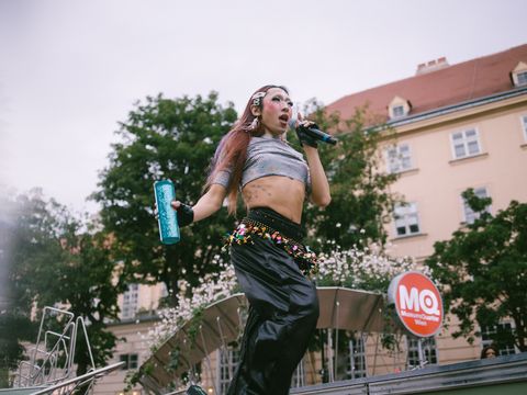 Person mit langen Haaren und bauchfreiem Oberteil hält Mikrofon und Trinkflasche auf einer Bühne vor einem Gebäude mit MQ-Logo