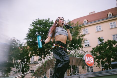 Person mit langen Haaren und bauchfreiem Oberteil hält Mikrofon und Trinkflasche auf einer Bühne vor einem Gebäude mit MQ-Logo