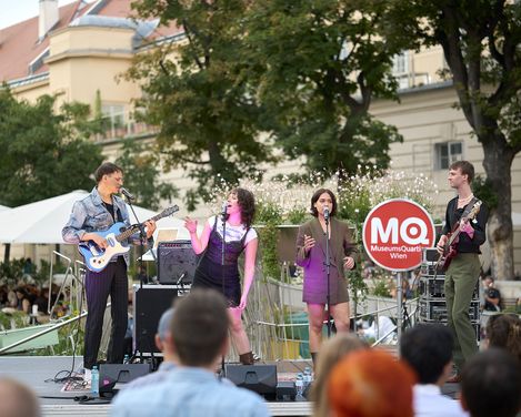 MQ Tunes, band: Low Life Rich Kids. Four people singing on stage in the MQ courtyard.
