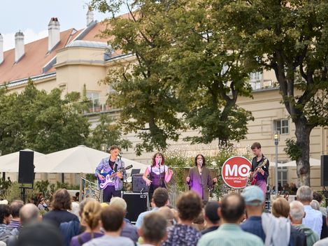 Mehrere Musiker mit Instrumenten und Mikrofonen stehen auf einer Bühne vor einer Menschenmenge im Freien, im Hintergrund Bäume und ein Gebäude mit roten Dachziegeln.
